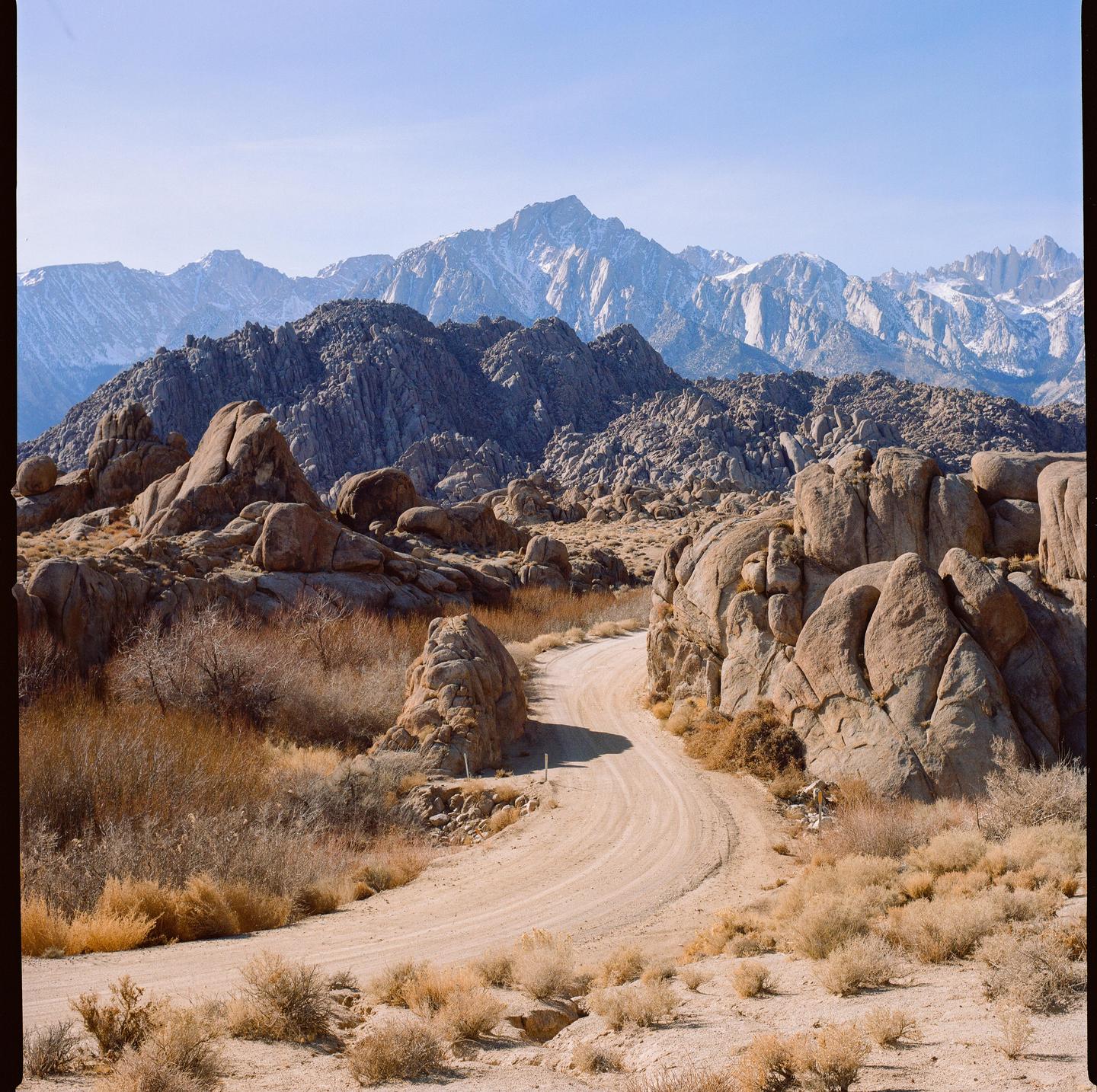 A winding dirt road leads through a rocky landscape towards snow-capped mountains under a clear sky.
