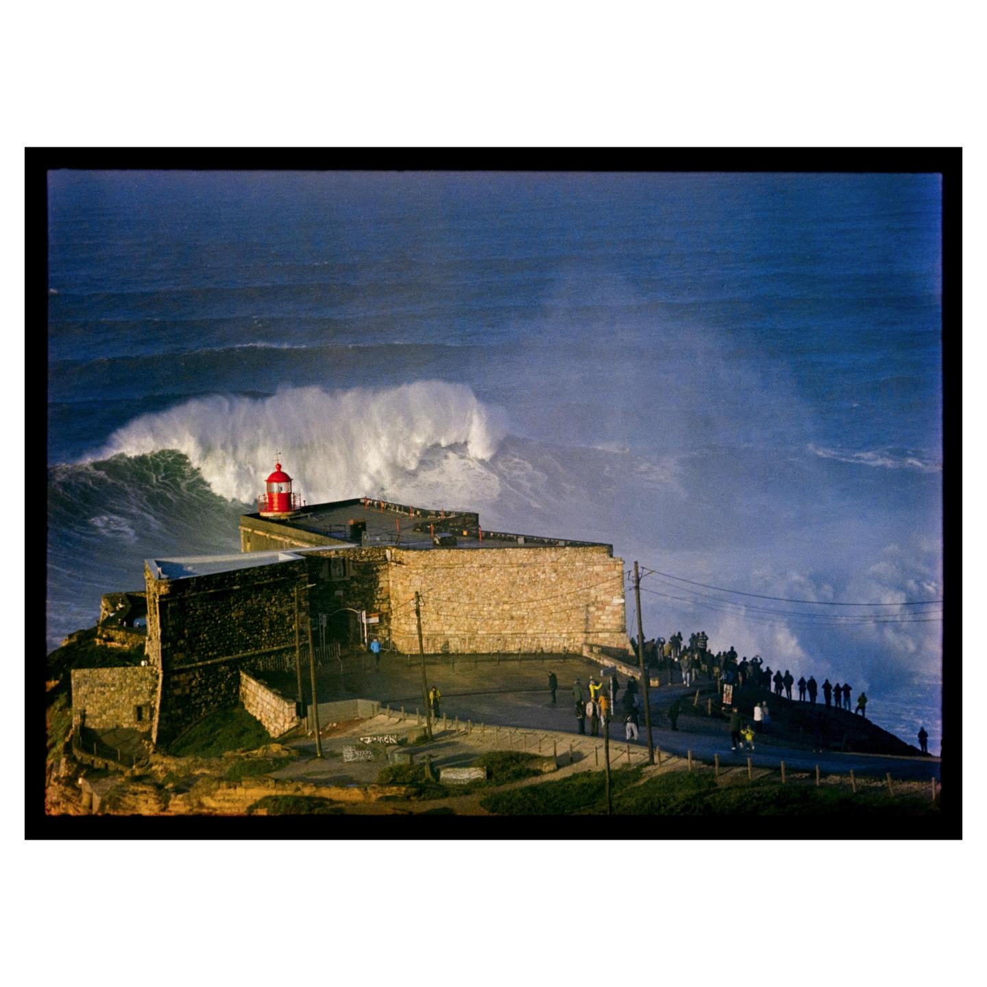 A large wave crashes behind a lighthouse and a stone building on a cliff where many people are gathered.