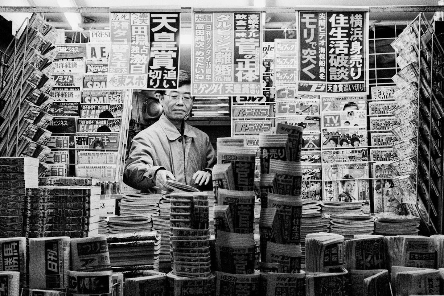 A man stands behind a counter filled with stacks of newspapers and magazines.