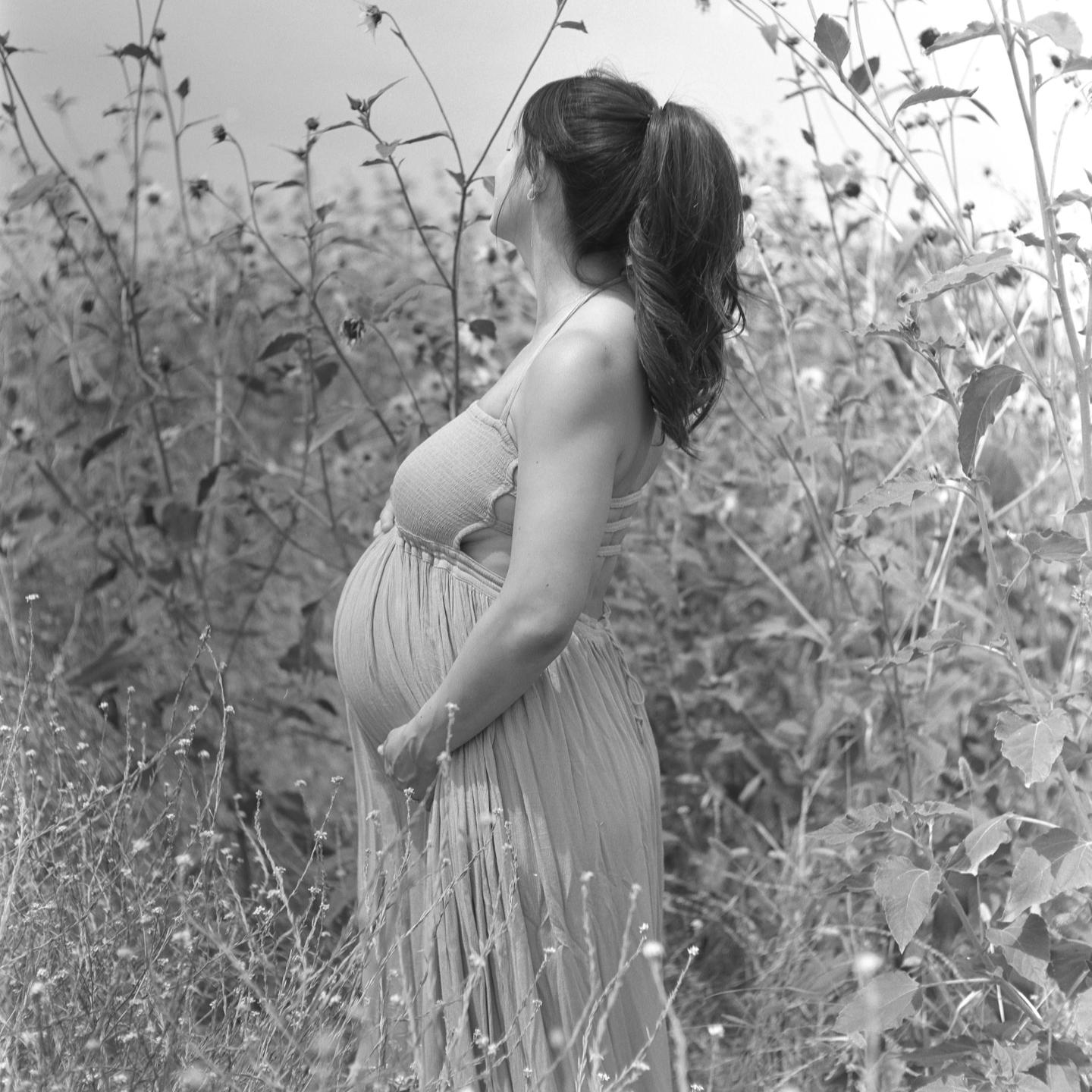 A pregnant woman stands in a field of tall plants, looking up and away from the camera.