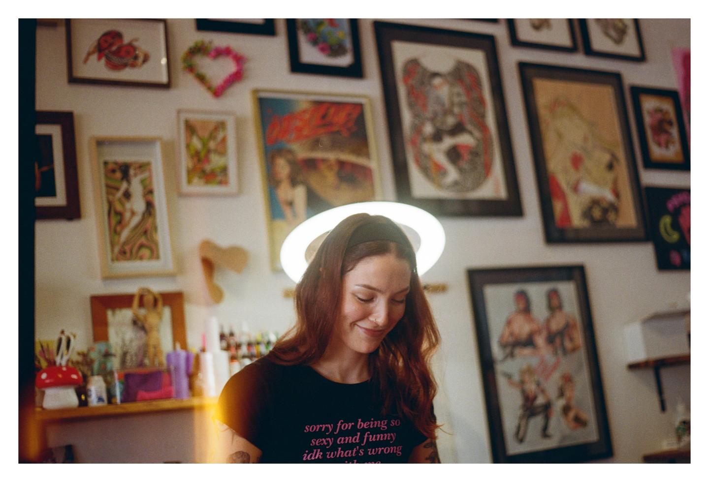 A woman with long hair and a black t-shirt stands in front of a wall covered with framed art.