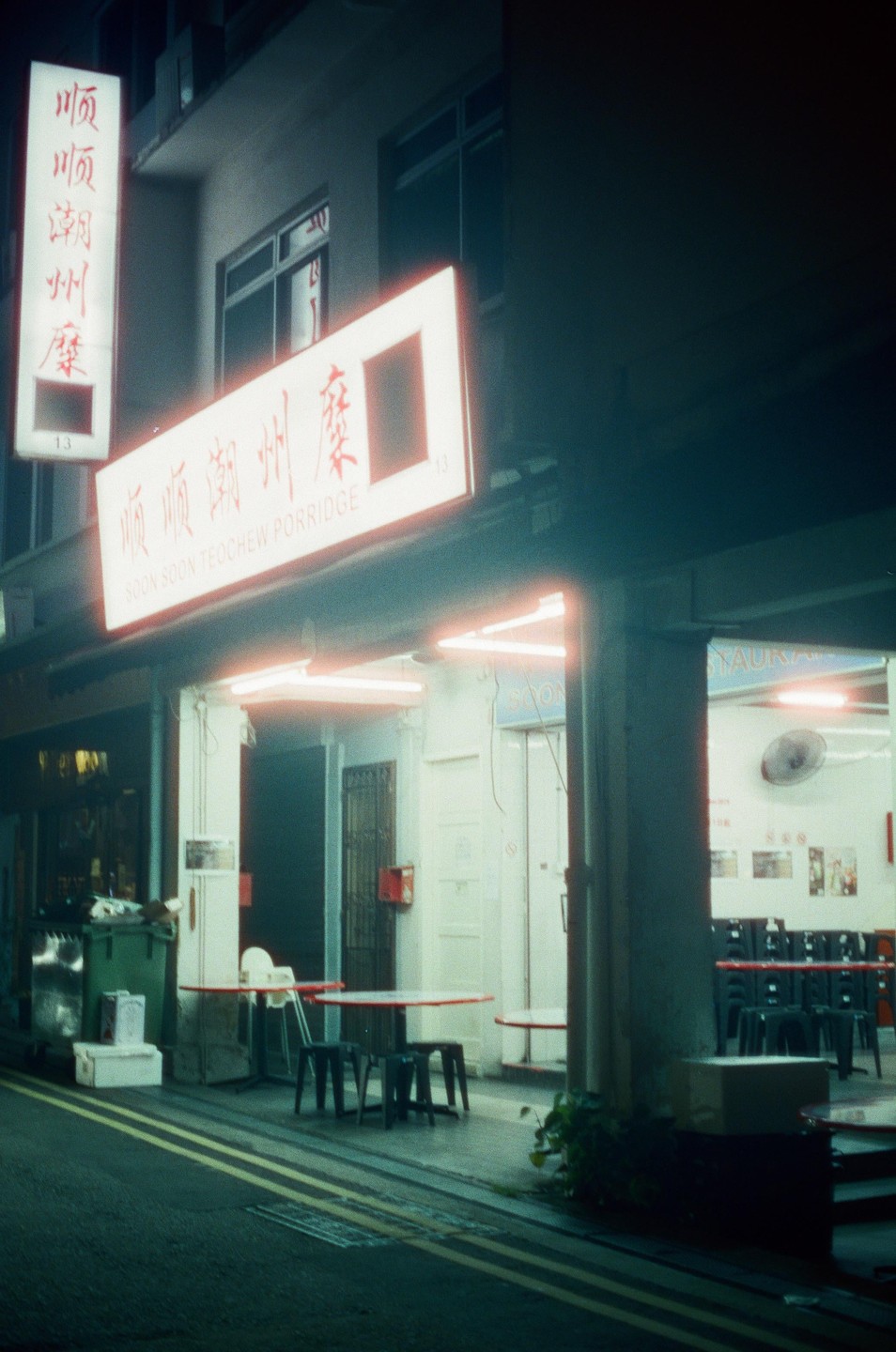 A street view of a restaurant with outdoor seating and neon signs at night.