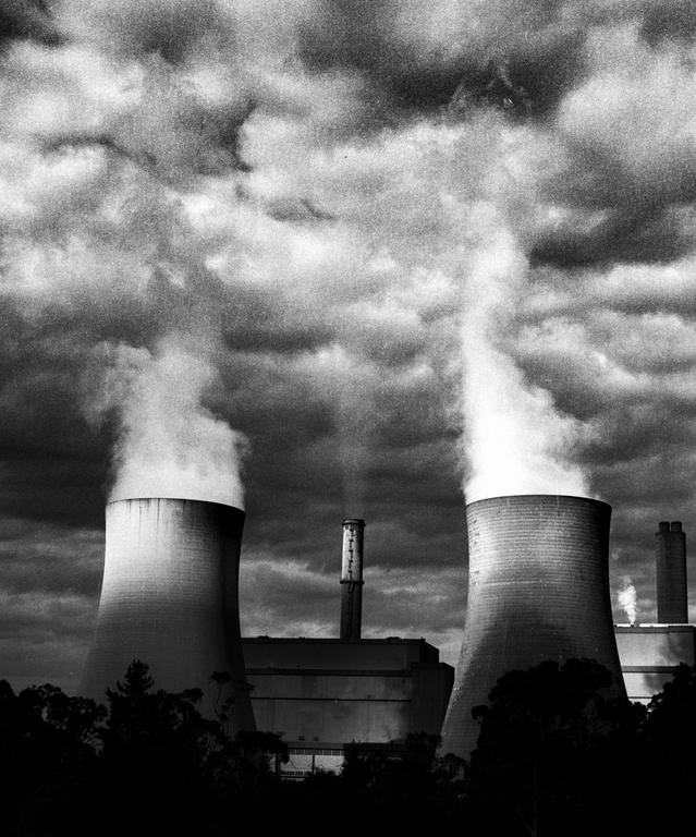 Two cooling towers emit large plumes of smoke into a cloudy sky above a power plant.