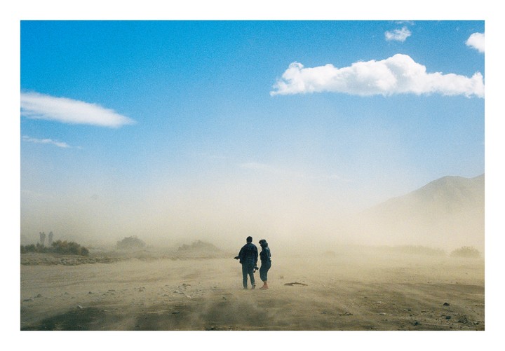 A couple stands in a desert landscape with a mountain in the background, surrounded by a dust storm.
