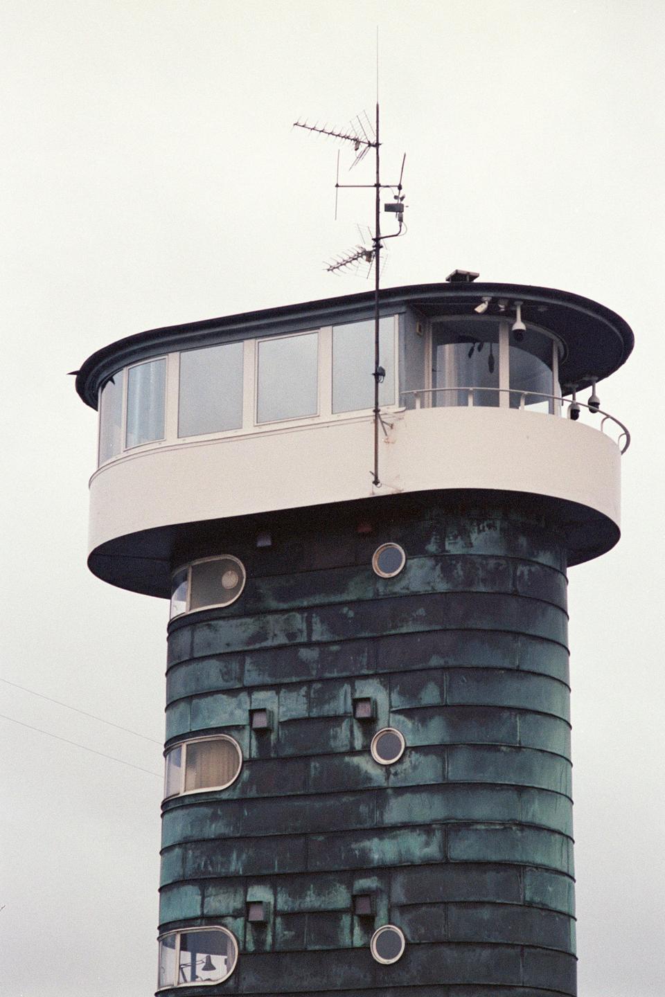 A tall, round building with a dark, textured facade and a lighter, windowed top section is visible against a plain sky.