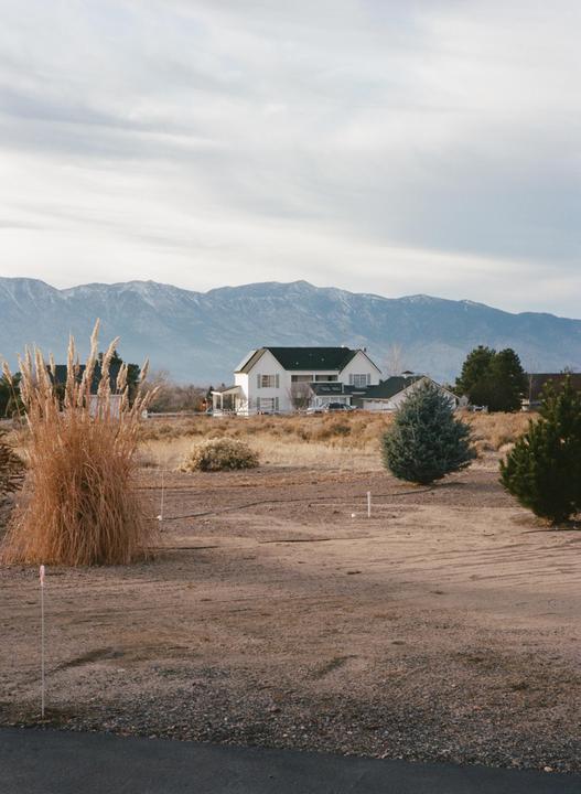 A white house with a dark roof stands in a dry, grassy field with mountains in the background.