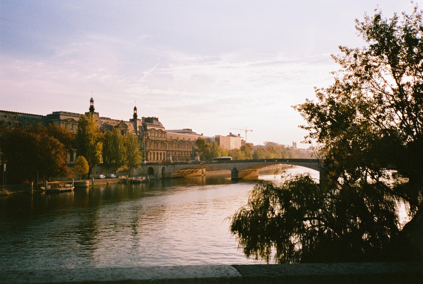 A river flows past a large building with turrets and a bridge, with trees in the foreground and background.