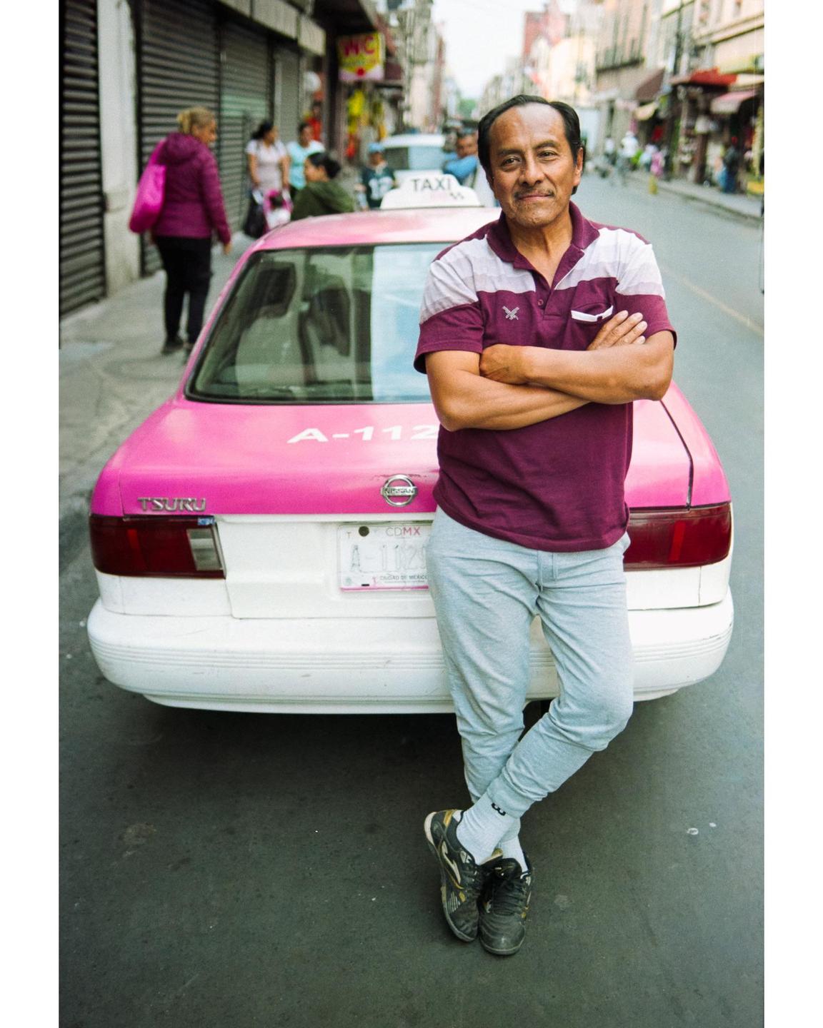 A man stands with his arms crossed, leaning against the back of a pink and white taxi on a city street.
