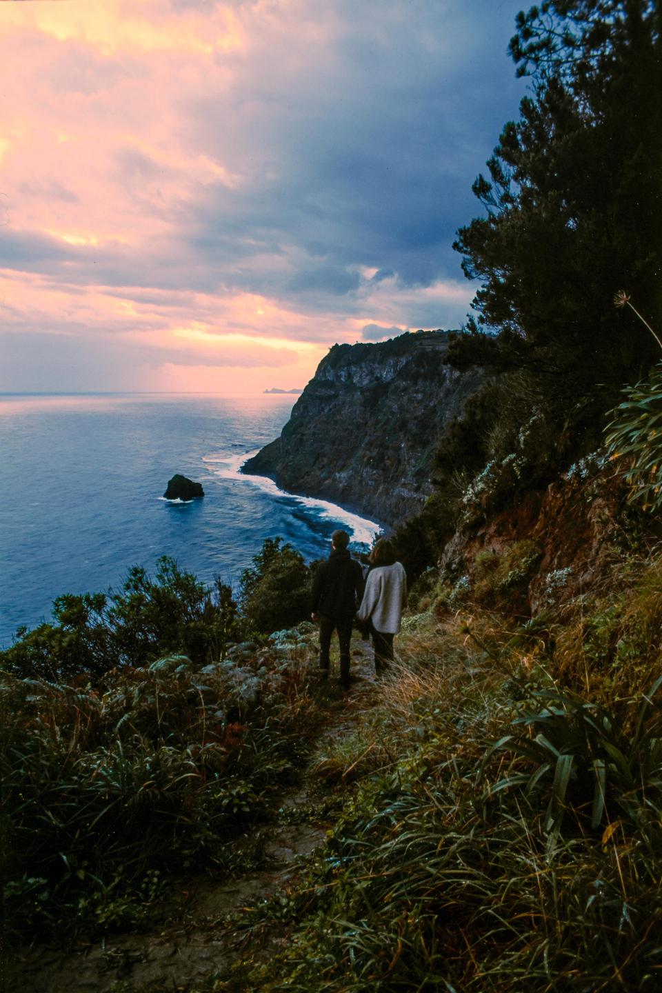 A couple stands on a cliffside path overlooking the ocean at sunset.