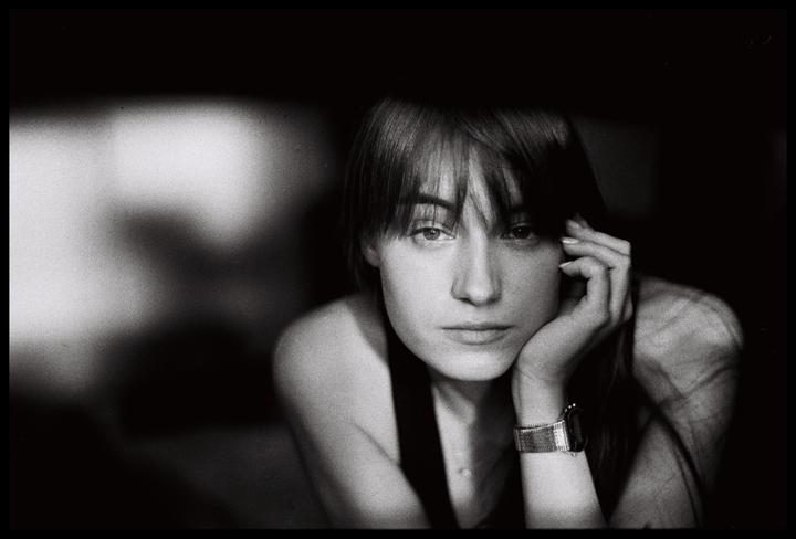 A woman with bangs and a watch rests her chin on her hand while looking at the viewer.