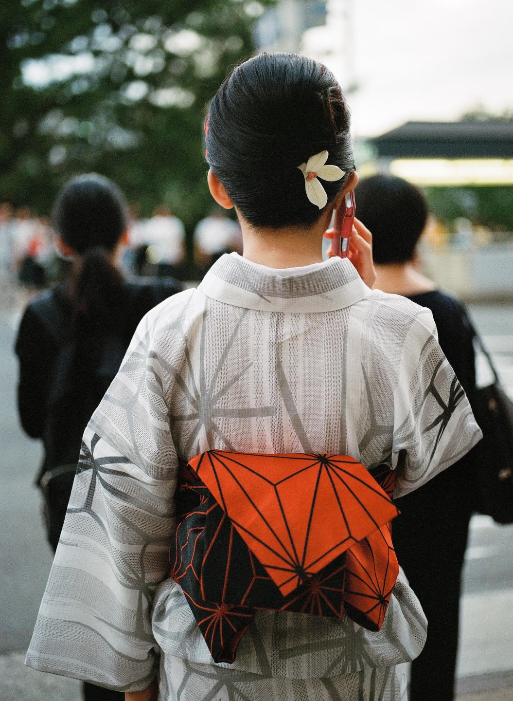 A woman in a kimono with an orange obi stands with her back to the viewer, holding a phone to her ear.