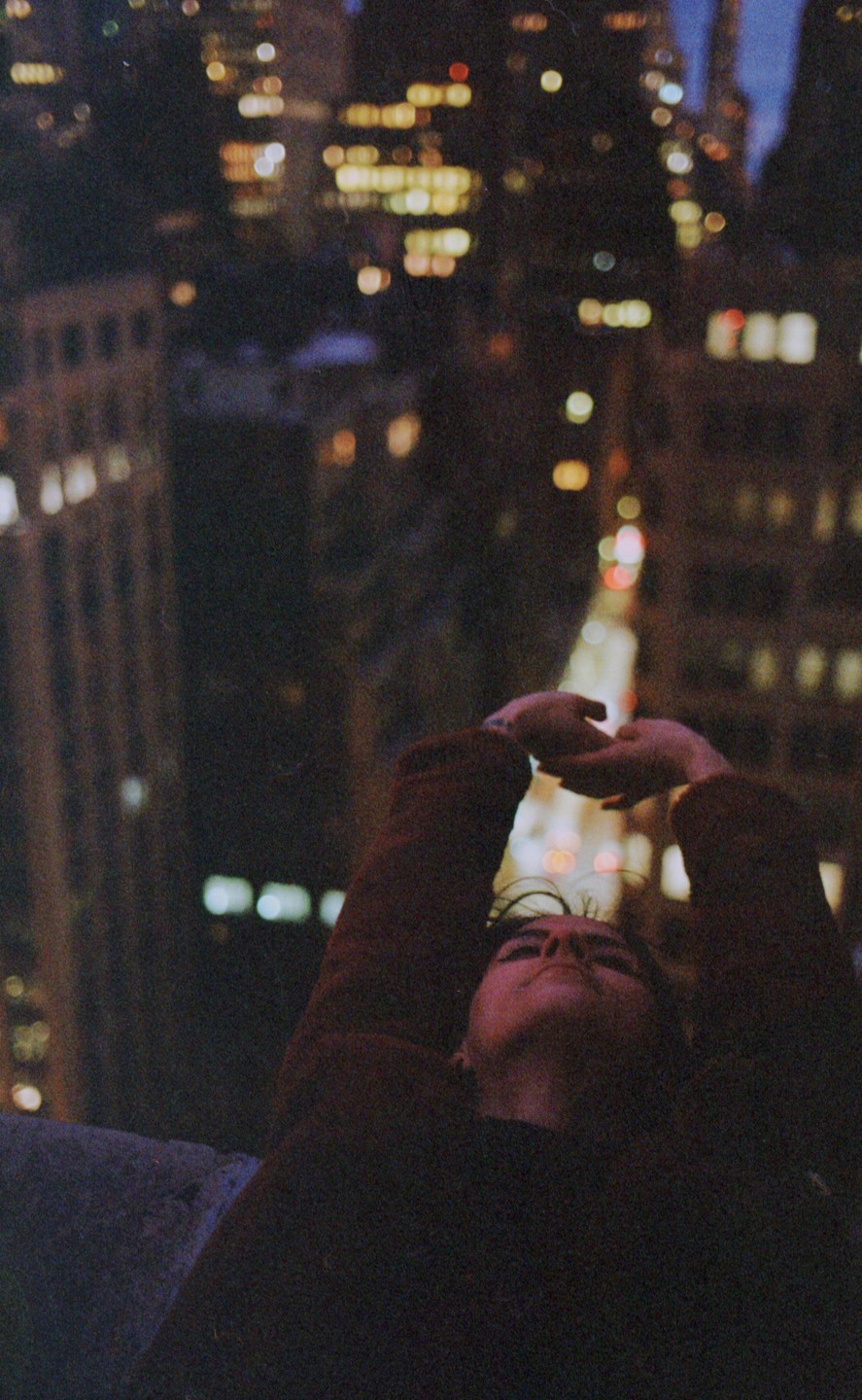 A person with arms raised looks up at the night sky above a city street with blurred lights.