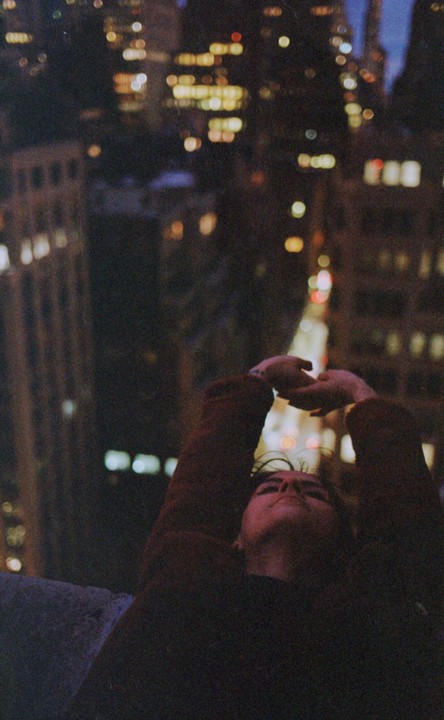 A person with arms raised looks up at the night sky above a city street with blurred lights.