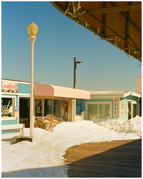 A boardwalk with snow drifts in front of several small shops under a clear blue sky.