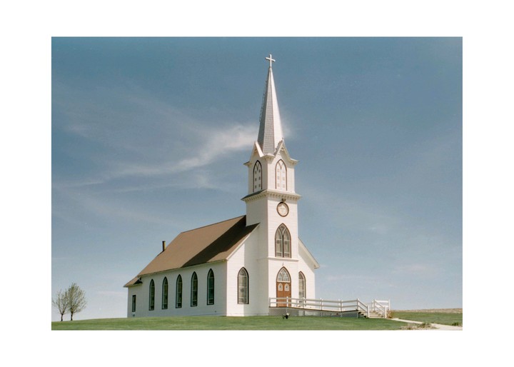 A white church with a tall steeple stands on a grassy hill under a blue sky.