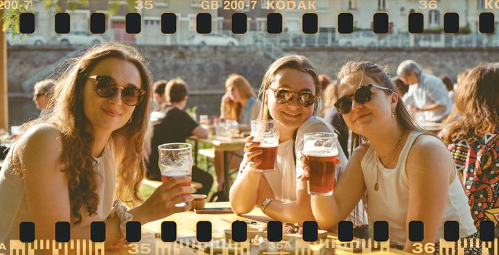 Three women wearing sunglasses are sitting at a table with drinks, smiling at the camera.