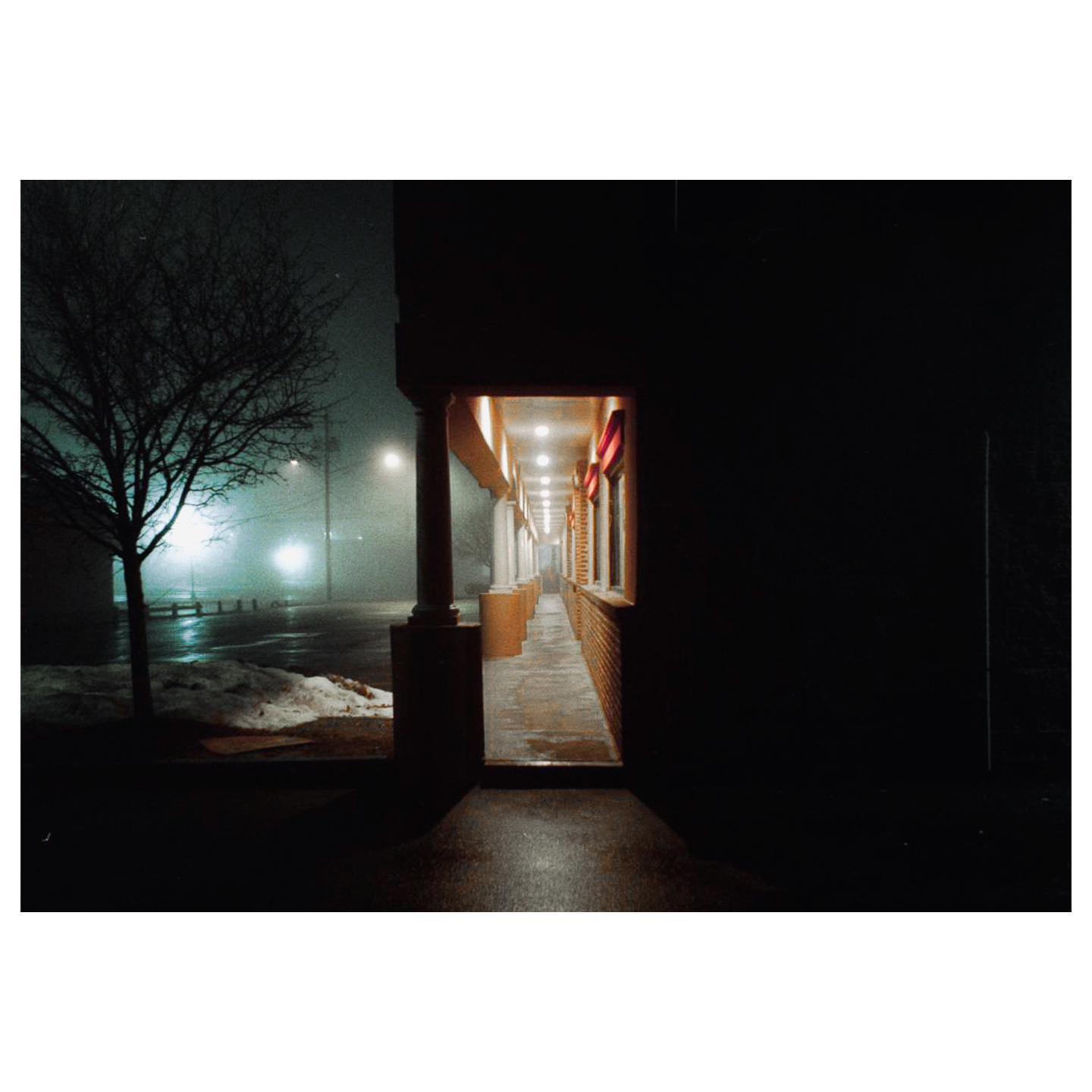 A tree stands next to a building with a long, illuminated walkway on a foggy night.