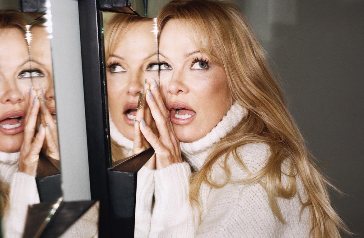 A woman with long blonde hair and a white turtleneck sweater looks into a mirror, with her reflection visible.
