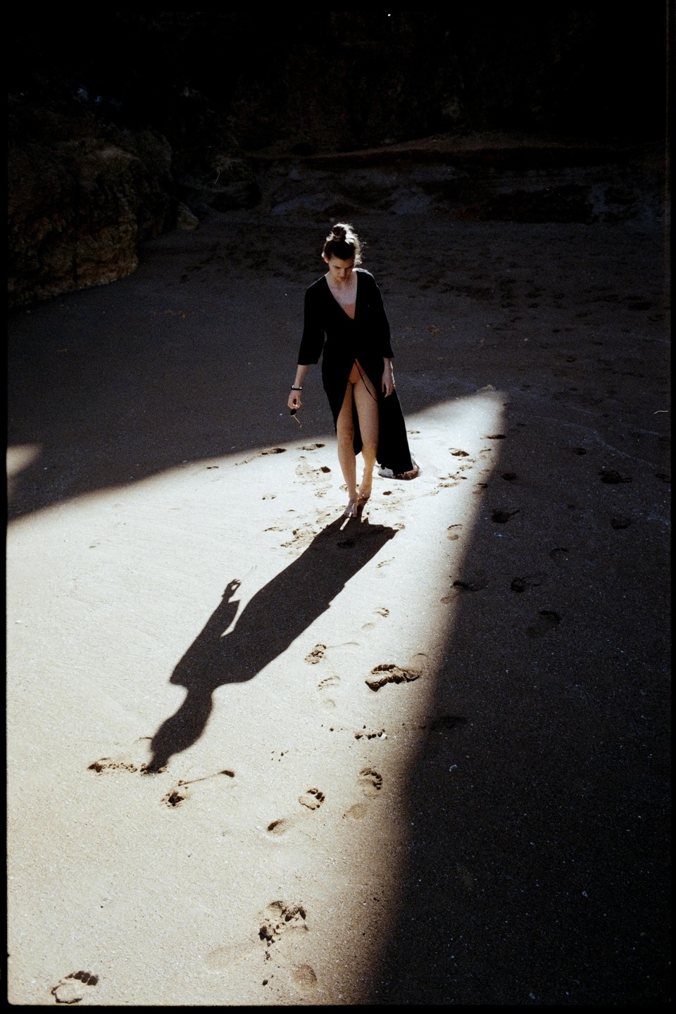 A woman walks barefoot on a sandy beach, casting a long shadow.