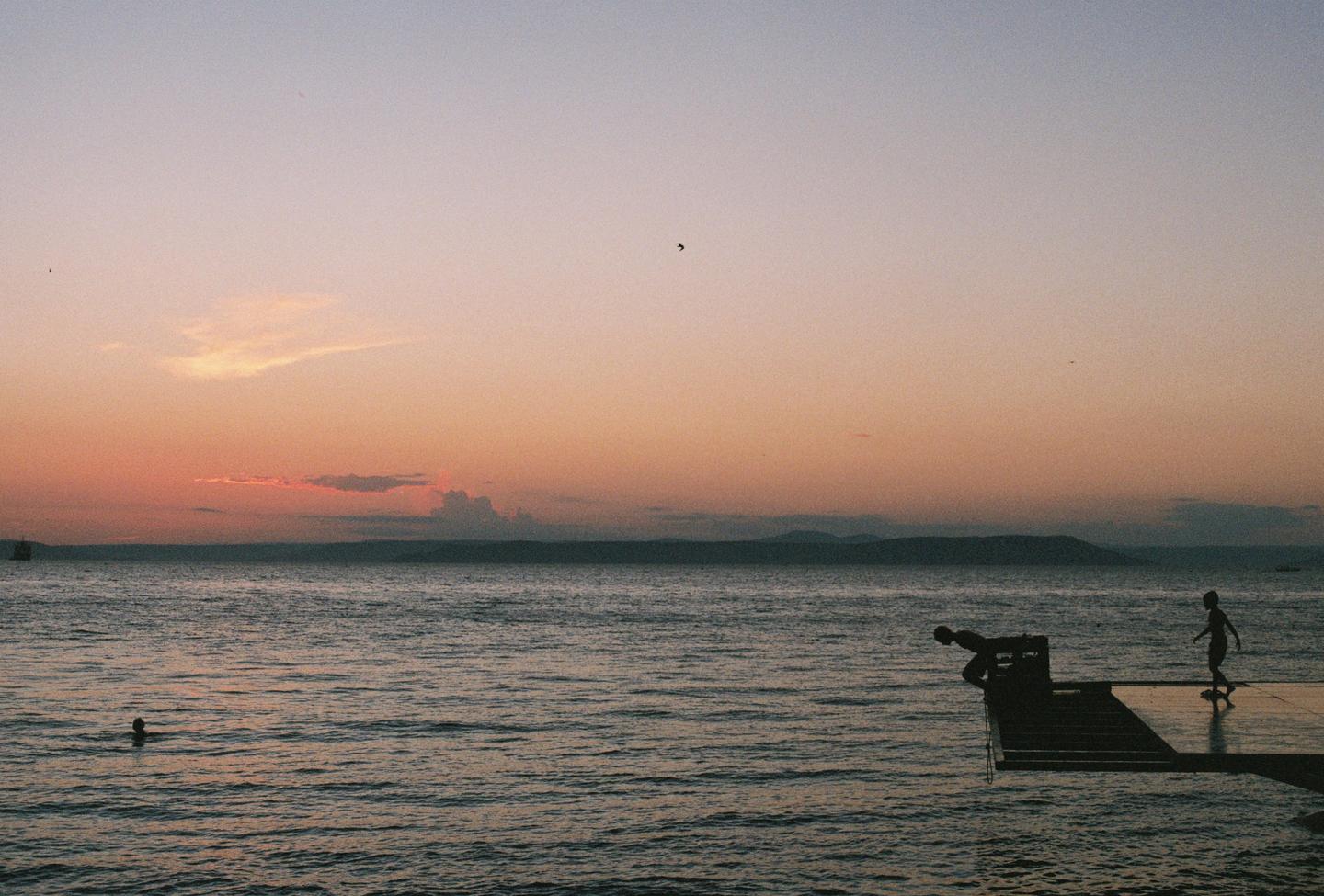 Two children are on a pier at sunset, with one child preparing to dive into the water and another standing nearby.