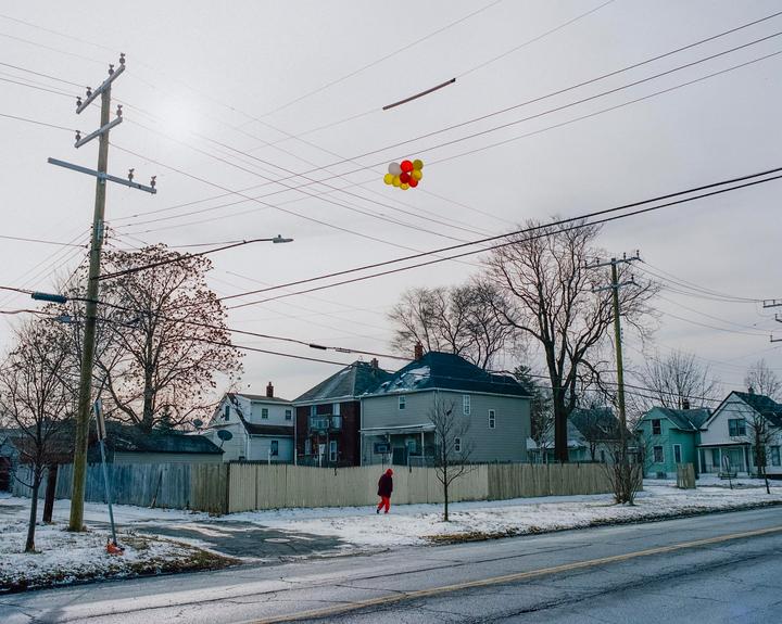 A person in a red coat walks along a snowy street with houses and bare trees, while balloons are tangled in overhead power lines.