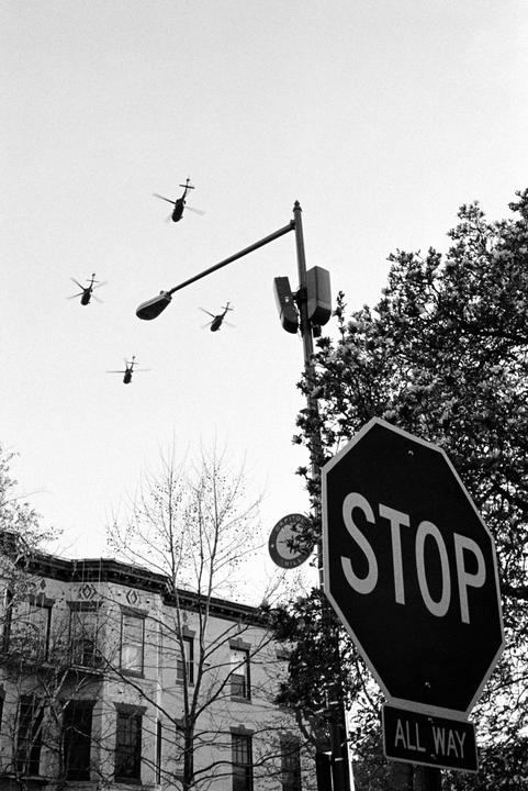 Four helicopters fly in the sky above a stop sign, a street light, trees, and a building.