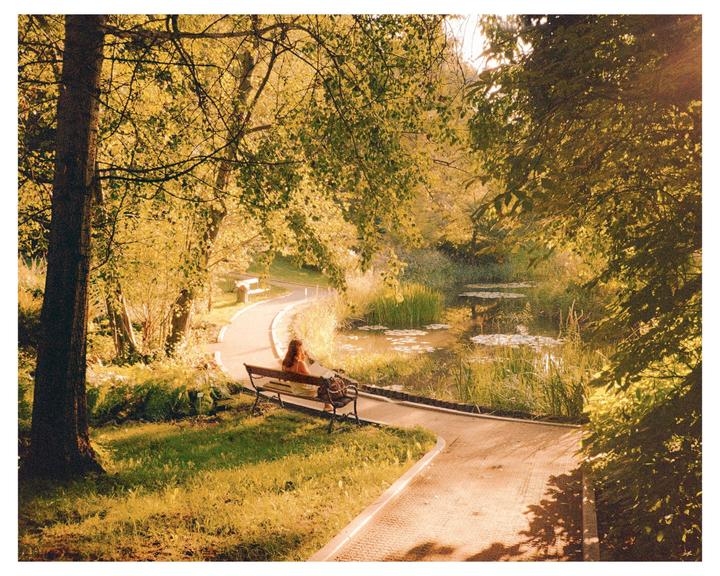 A woman sits on a bench next to a winding path and a pond surrounded by trees with autumn foliage.