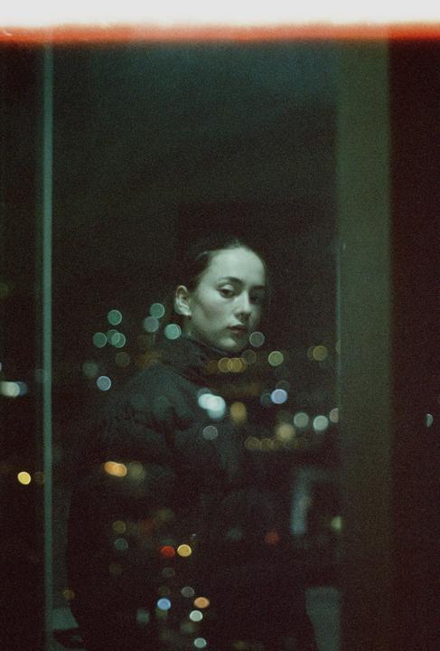 A woman stands in front of a window at night, with city lights visible as bokeh in the background.