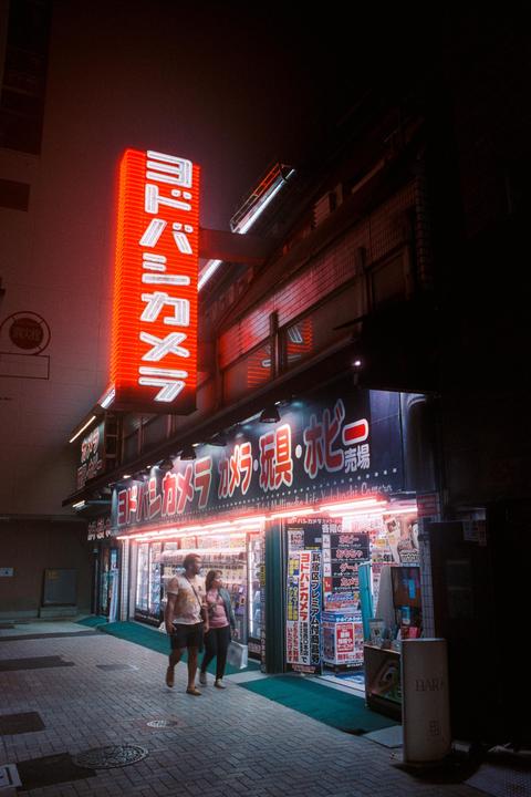 A couple walks past a brightly lit storefront with neon signs at night.
