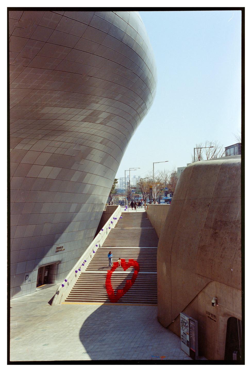 A large, modern building with a curved facade stands next to a wide staircase with a red heart sculpture at its base.