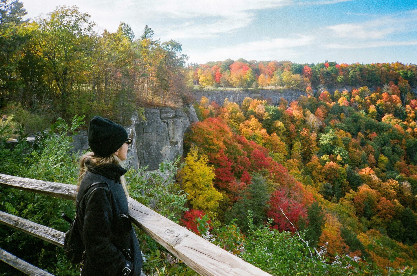 A woman in a black coat and hat stands on a wooden railing overlooking a valley filled with autumn foliage.