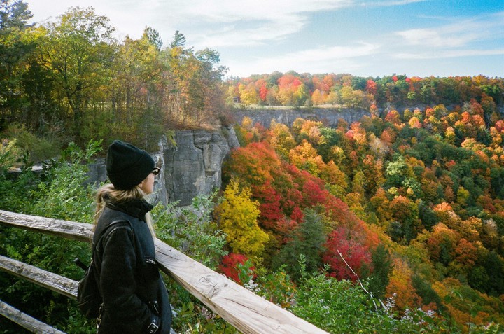 A woman in a black coat and hat stands on a wooden railing overlooking a valley filled with autumn foliage.