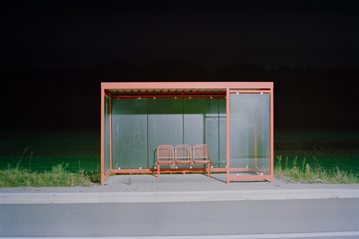 A bus stop with three red seats stands on the side of a road at night, with a dark field and trees in the background.
