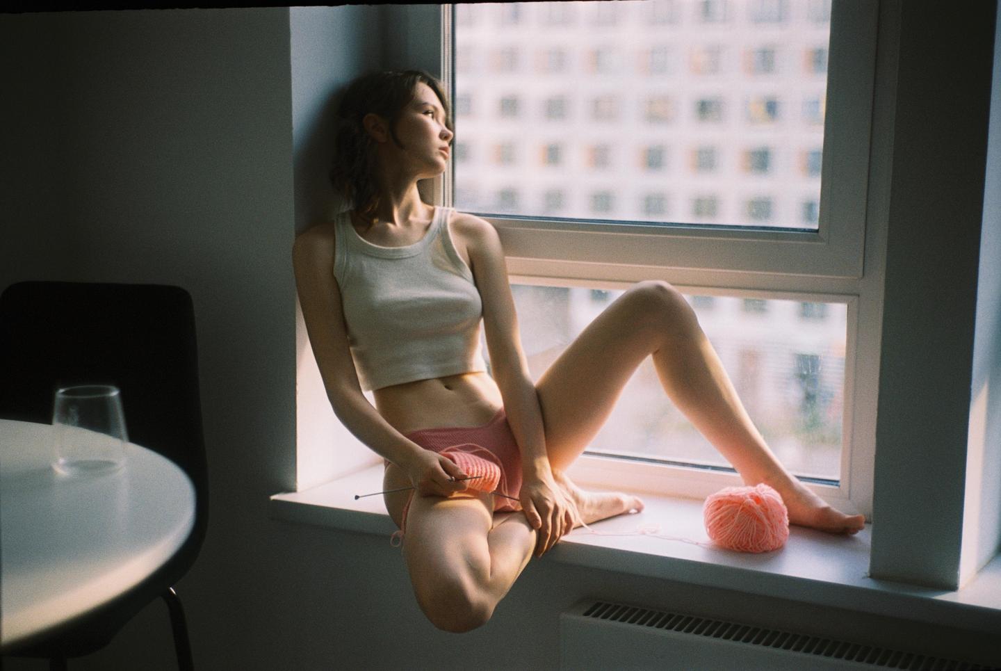 A woman sits on a windowsill knitting with pink yarn.