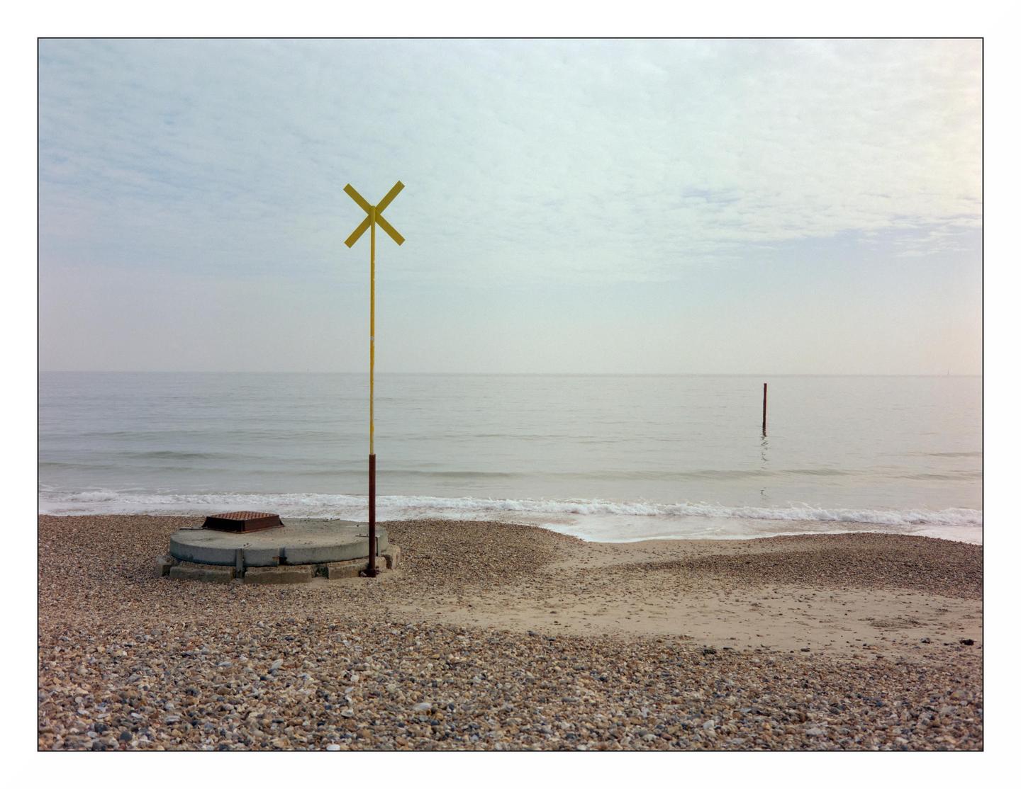 A yellow marker with an X shape stands on a concrete base on a pebble beach next to the ocean under a cloudy sky.