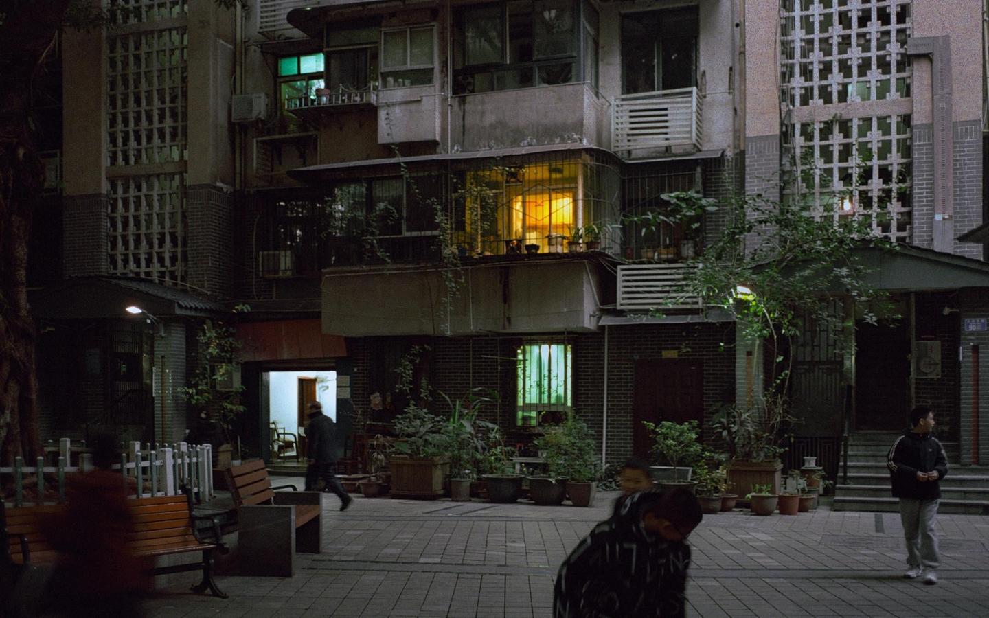 A street at night is lined with buildings and potted plants, with people walking and a bright yellow light emanating from a window.