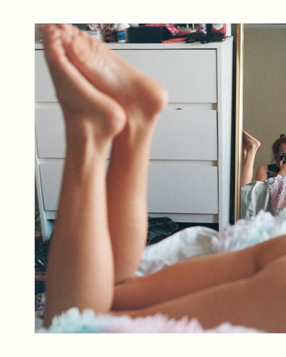 A person's legs are visible in the foreground with their feet raised, and a mirror reflection of a woman holding a camera is visible in the background.