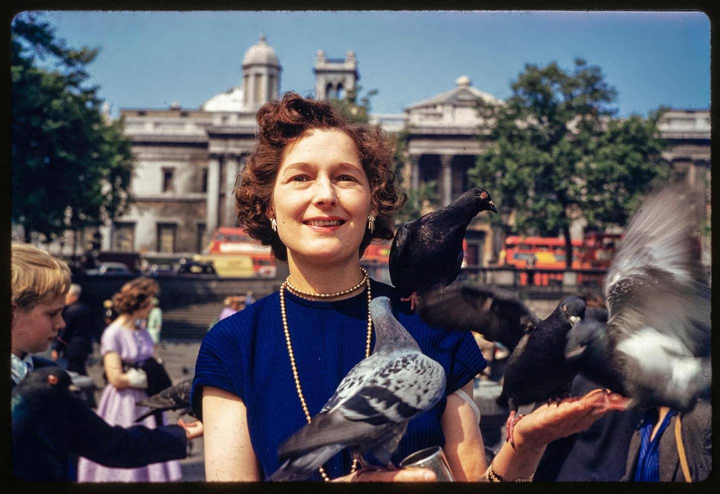 A smiling woman with pigeons on her shoulders and arm stands in a city square with a large building in the background.