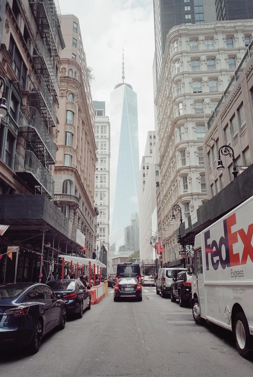 A street in a city is lined with parked cars and tall buildings, with a skyscraper visible in the distance.