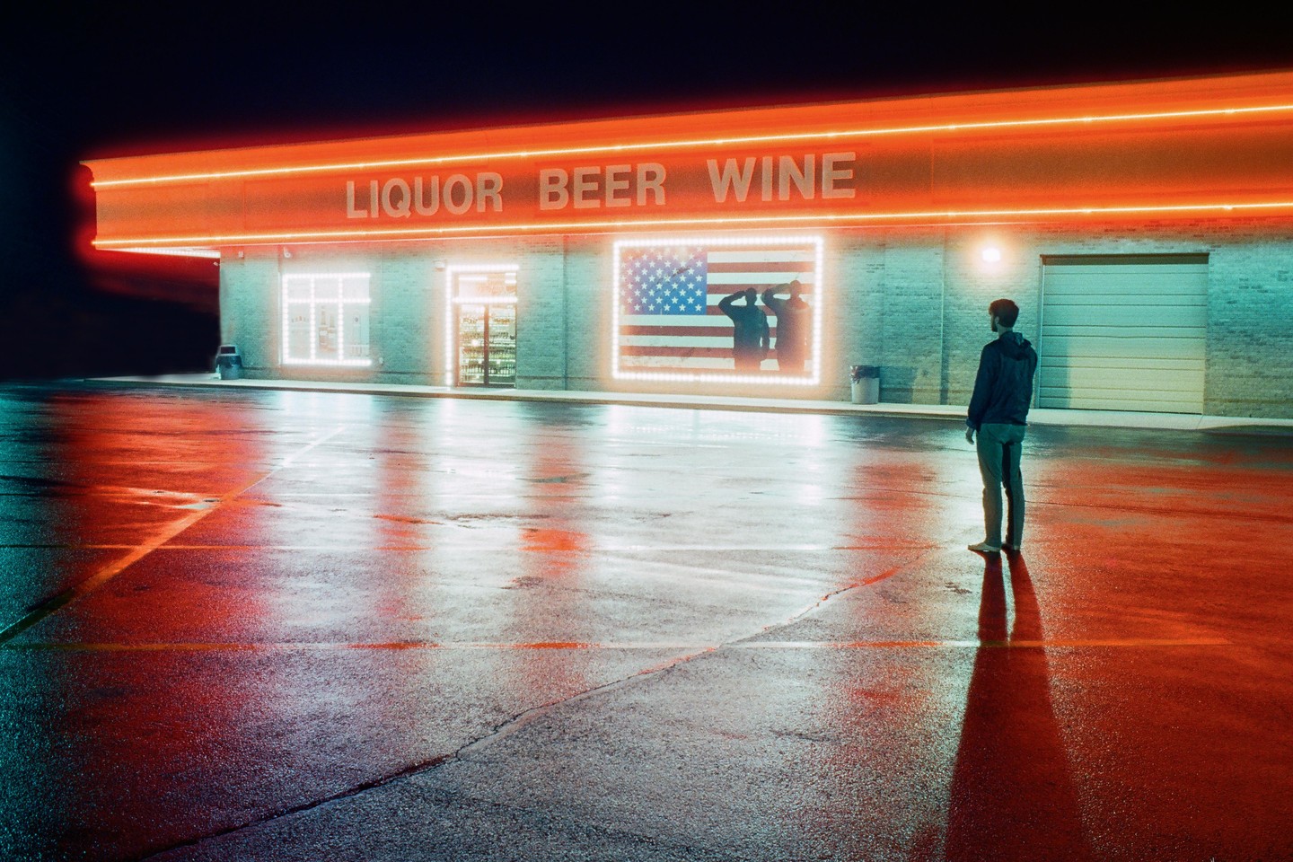 A man stands in a wet parking lot in front of a liquor store with a neon American flag sign at night.