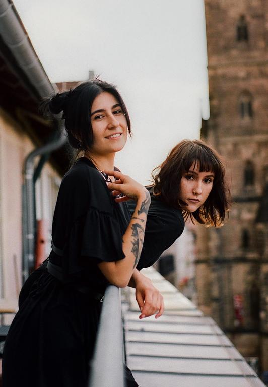Two women lean over a balcony railing with a building in the background.