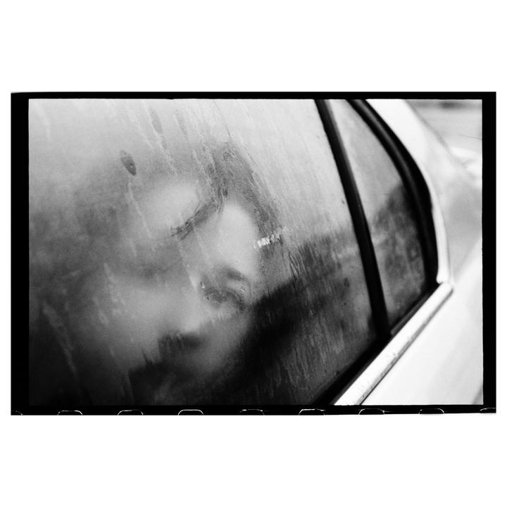 A person's face is visible through a wet car window.