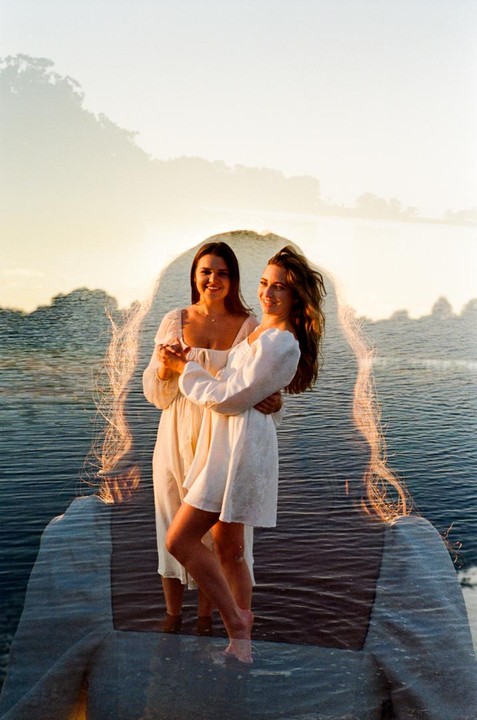Two women in white dresses stand in water at sunset, with a double exposure of a woman's silhouette overlaid.