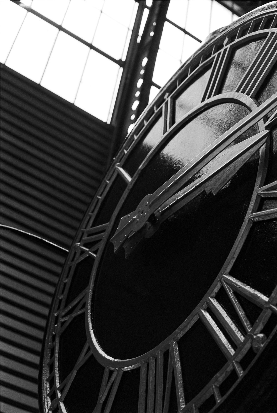 A large clock face with roman numerals and hands is seen from a low angle, with a glass ceiling in the background.
