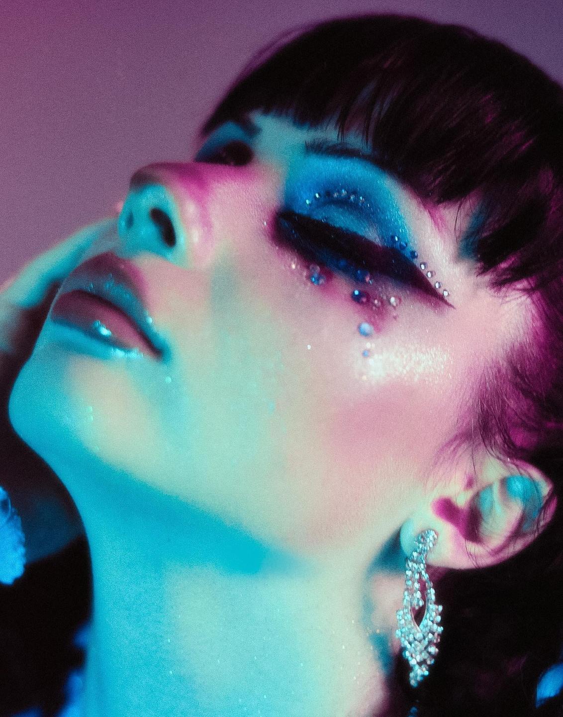 A woman with dramatic blue and glittery eye makeup and a large earring is illuminated by colorful light.