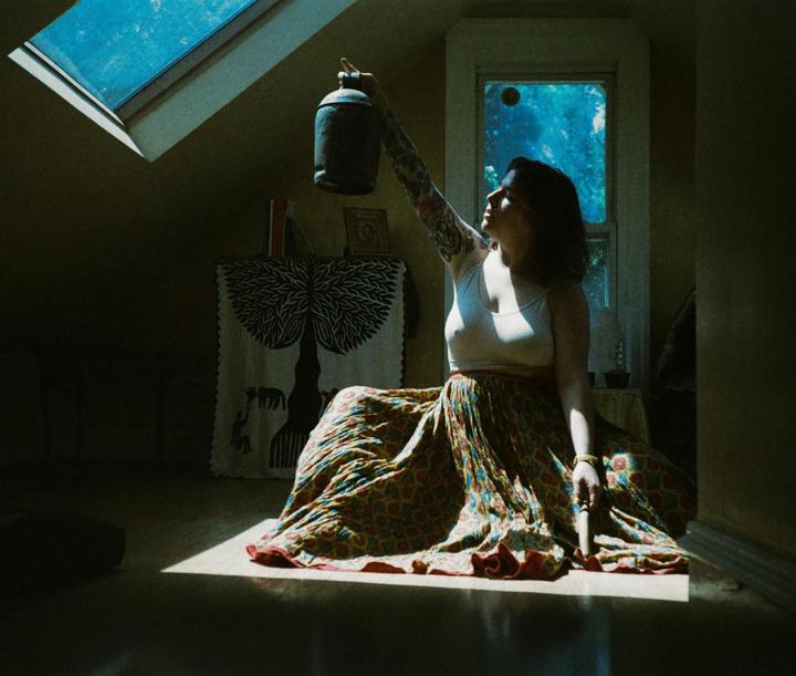 A woman sits on the floor in a sunlit room, holding a lantern up towards a skylight.