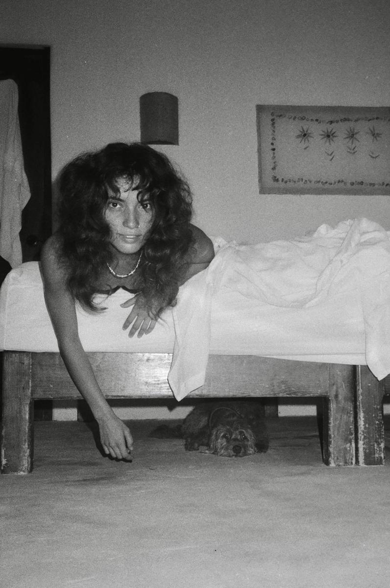 A woman with long curly hair lies on a bed, looking at the camera, while a small dog peeks out from underneath the bed.
