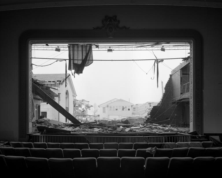 The interior of a damaged theater with rows of seats faces a stage opening that reveals a destroyed town.