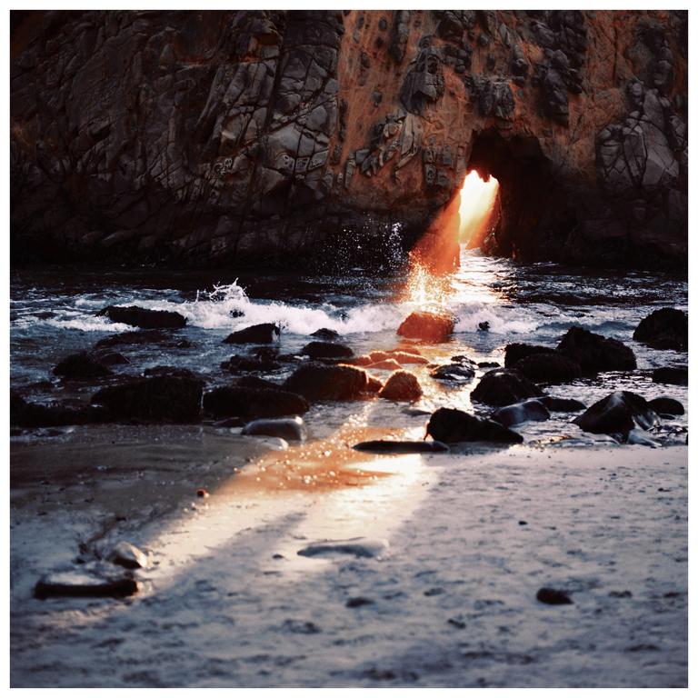 Sunlight streams through a rock arch onto a sandy beach with waves crashing over rocks.