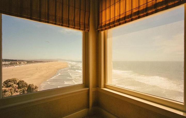 A view of a beach and ocean is seen through two windows with blinds.